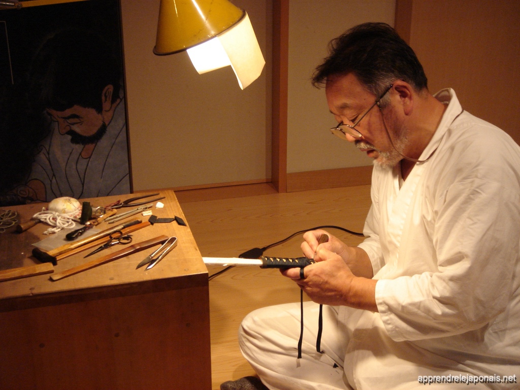 Artisan en tenue blanche tressant la poignée d'un katana, ses outils posés sur une table