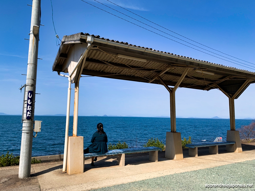 Une femme assise sur un banc du quai de la gare de Shimonada, face à la mer intérieure de Seto